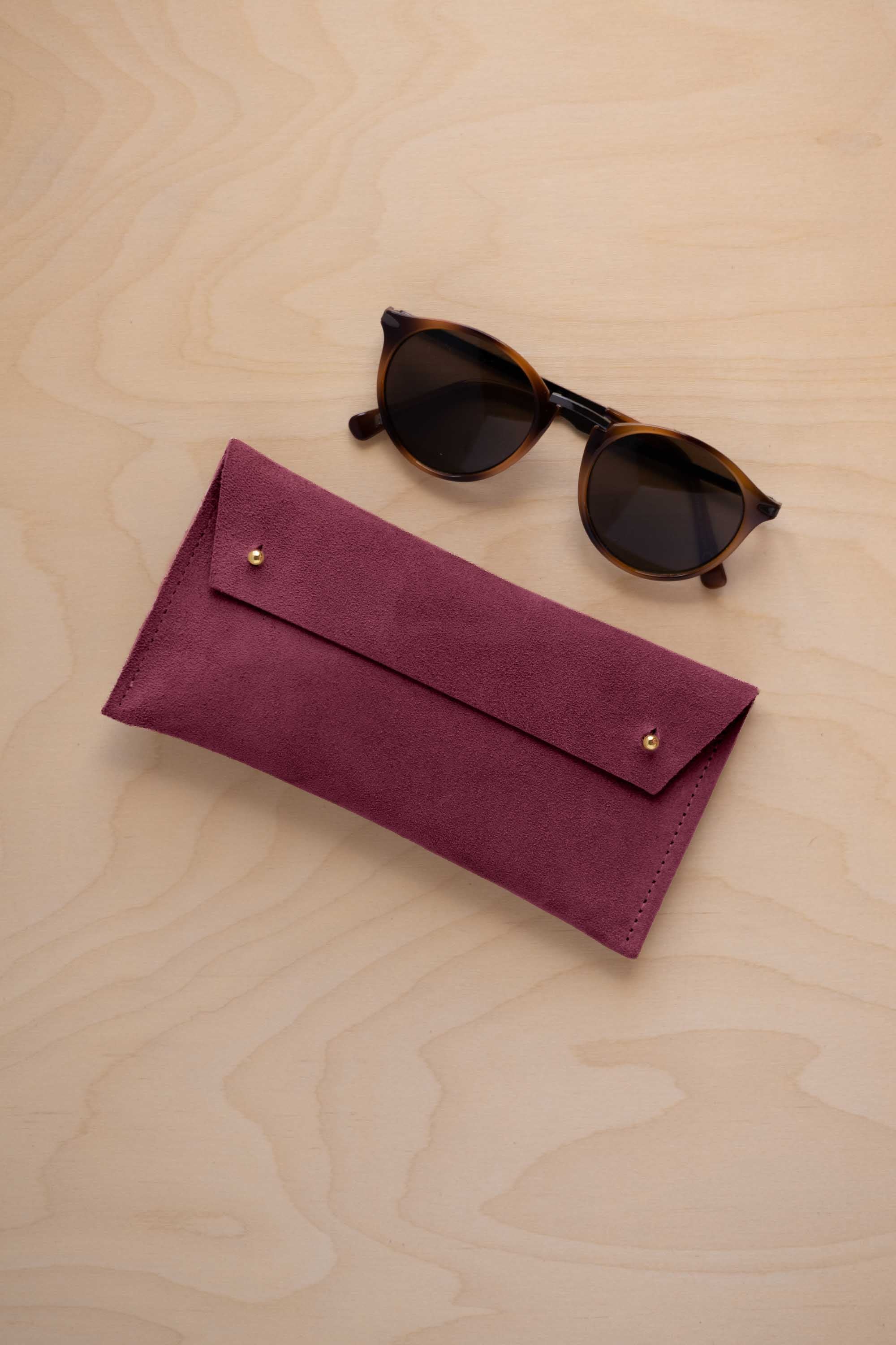 A pair of dark brown tortoiseshell sunglasses with round frames, resting on a light beige wood-grain surface next to a burgundy suede glasses case with gold coloured buttons.