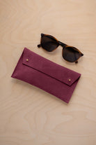 A pair of dark brown tortoiseshell sunglasses with round frames, resting on a light beige wood-grain surface next to a burgundy suede glasses case with gold coloured buttons.