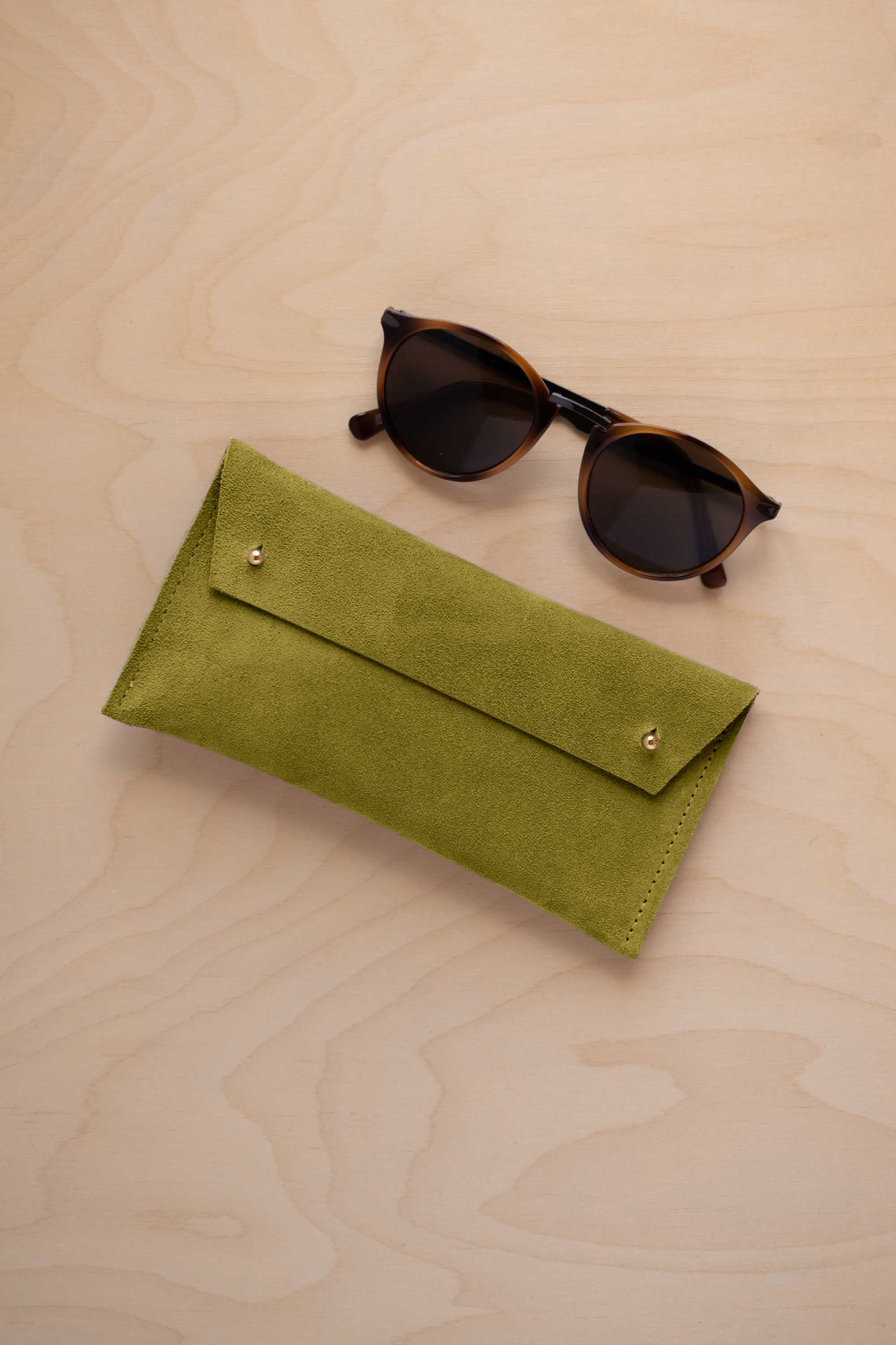 A pair of dark brown tortoiseshell sunglasses with round frames, resting on a light beige wood-grain surface next to a chartreuse lime green coloured suede glasses case with gold coloured buttons.
