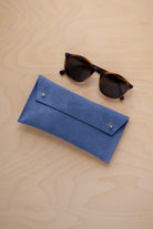 A pair of dark brown tortoiseshell sunglasses with round frames, resting on a light beige wood-grain surface next to a cobalt blue suede glasses case with gold coloured buttons.