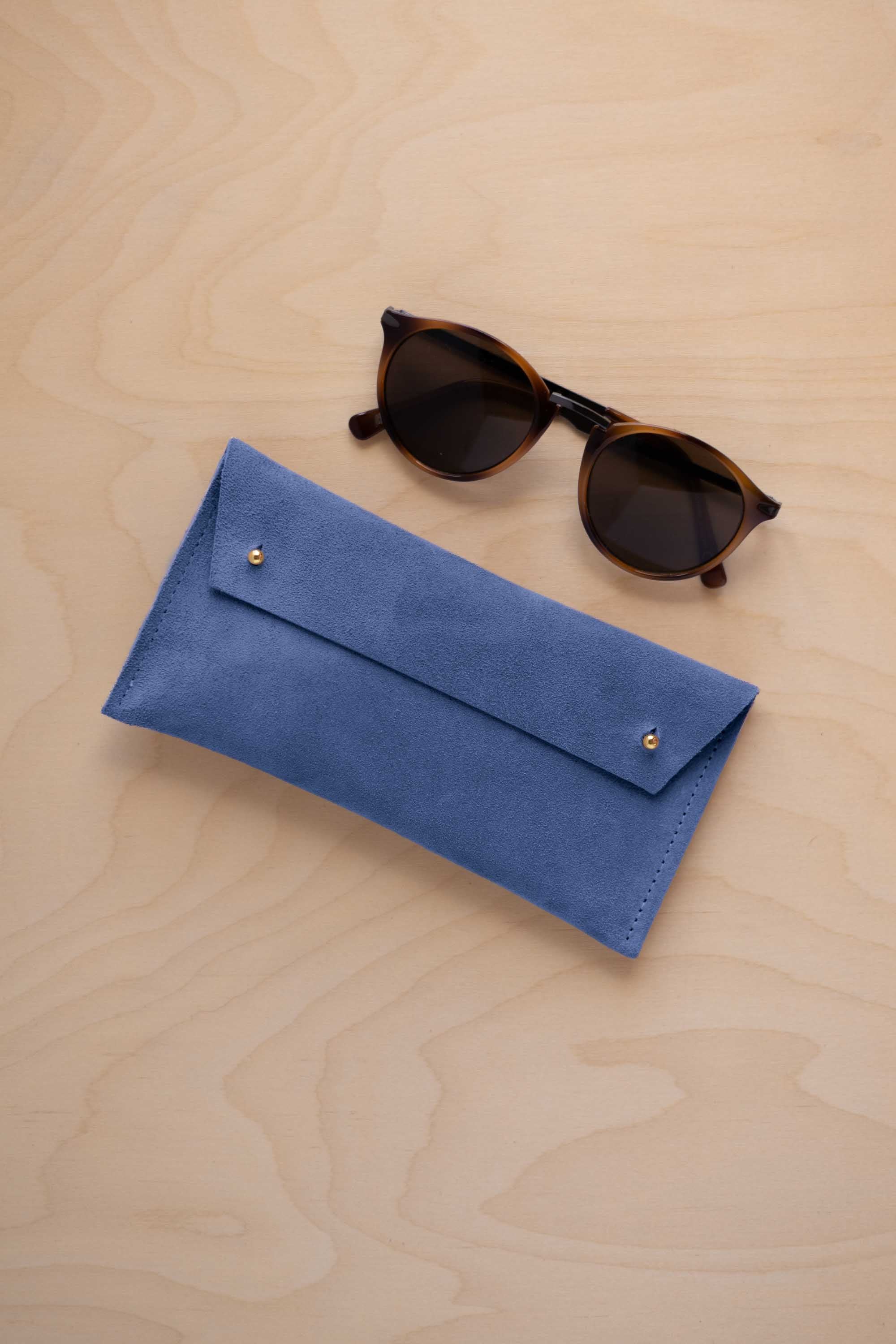 A pair of dark brown tortoiseshell sunglasses with round frames, resting on a light beige wood-grain surface next to a cobalt blue suede glasses case with gold coloured buttons.