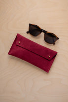 A pair of dark brown tortoiseshell sunglasses with round frames, resting on a light beige wood-grain surface next to a crimson red suede glasses case with gold coloured buttons.