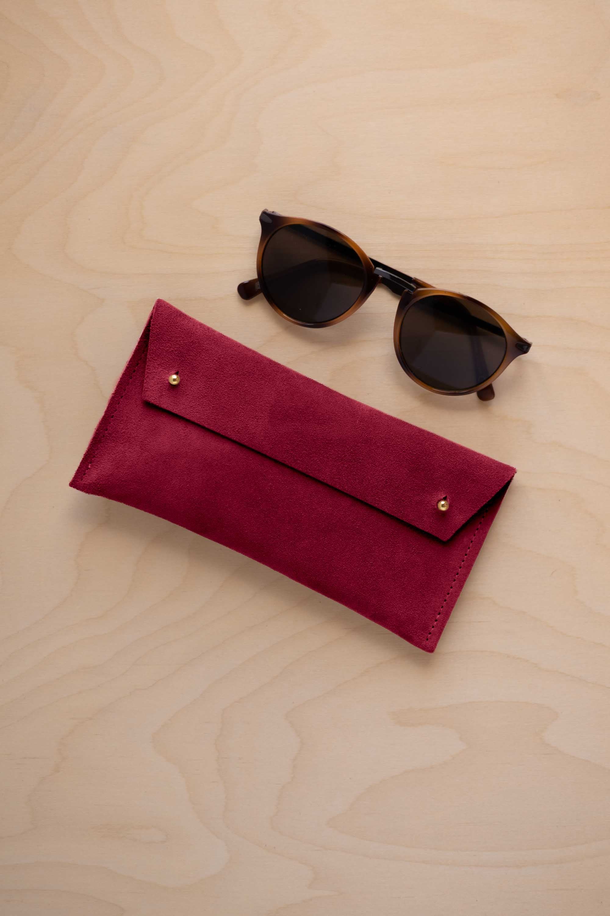 A pair of dark brown tortoiseshell sunglasses with round frames, resting on a light beige wood-grain surface next to a crimson red suede glasses case with gold coloured buttons.