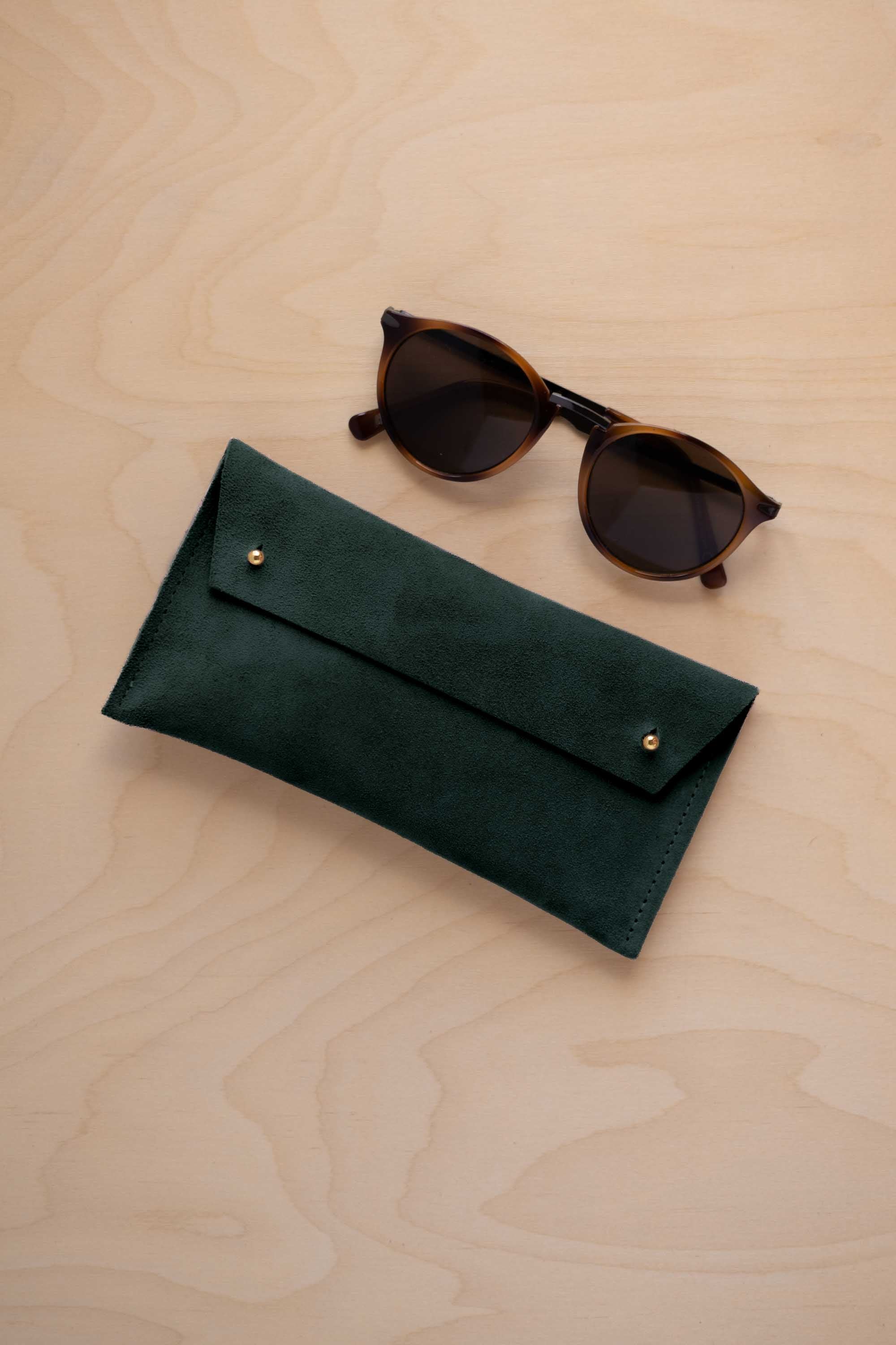 A pair of dark brown tortoiseshell sunglasses with round frames, resting on a light beige wood-grain surface next to a deep forest green suede glasses case with gold coloured buttons.