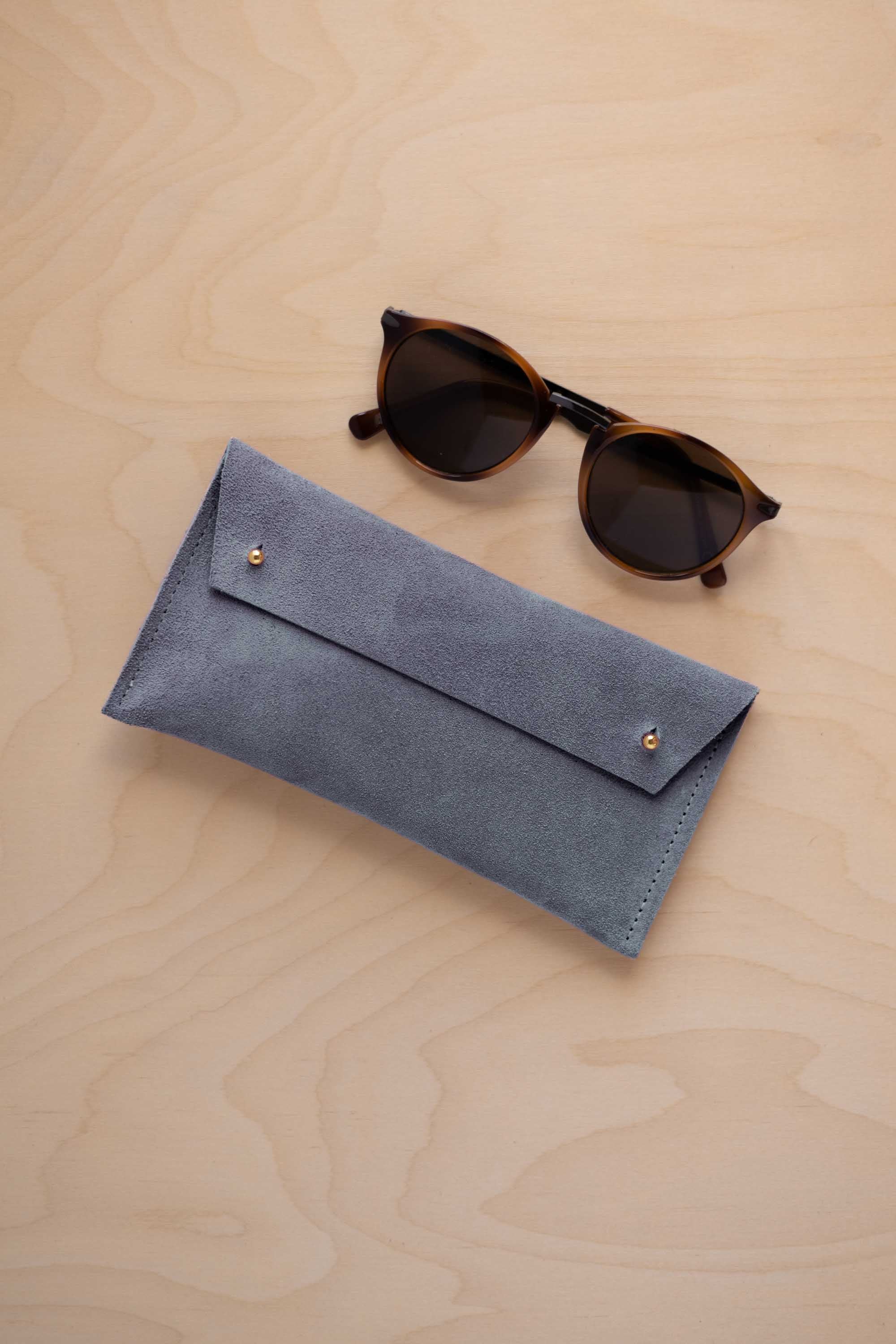 A pair of dark brown tortoiseshell sunglasses with round frames, resting on a light beige wood-grain surface next to a dusky blue suede glasses case with gold coloured buttons.