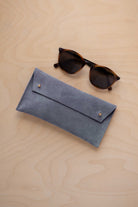 A pair of dark brown tortoiseshell sunglasses with round frames, resting on a light beige wood-grain surface next to a dusky blue suede glasses case with gold coloured buttons.