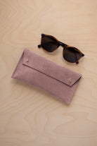 A pair of dark brown tortoiseshell sunglasses with round frames, resting on a light beige wood-grain surface next to a dusty mauve grey suede glasses case with gold coloured buttons.