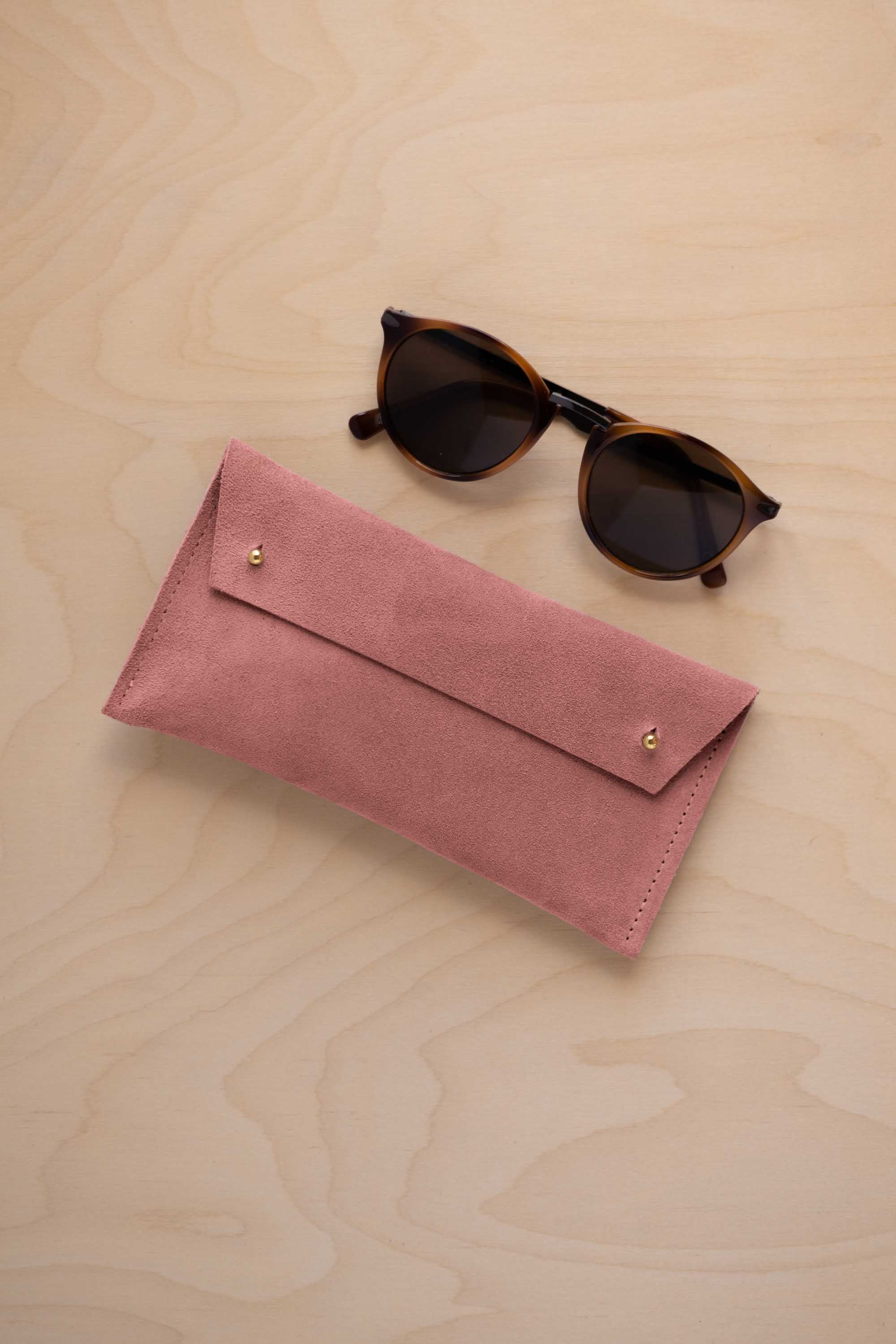 A pair of dark brown tortoiseshell sunglasses with round frames, resting on a light beige wood-grain surface next to a dusty rose pink coloured suede glasses case with gold coloured buttons.