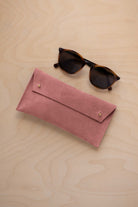 A pair of dark brown tortoiseshell sunglasses with round frames, resting on a light beige wood-grain surface next to a dusty rose pink coloured suede glasses case with gold coloured buttons.