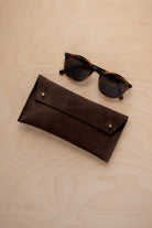 A pair of dark brown tortoiseshell sunglasses with round frames, resting on a light beige wood-grain surface next to an espresso brown coloured suede glasses case with gold coloured buttons.