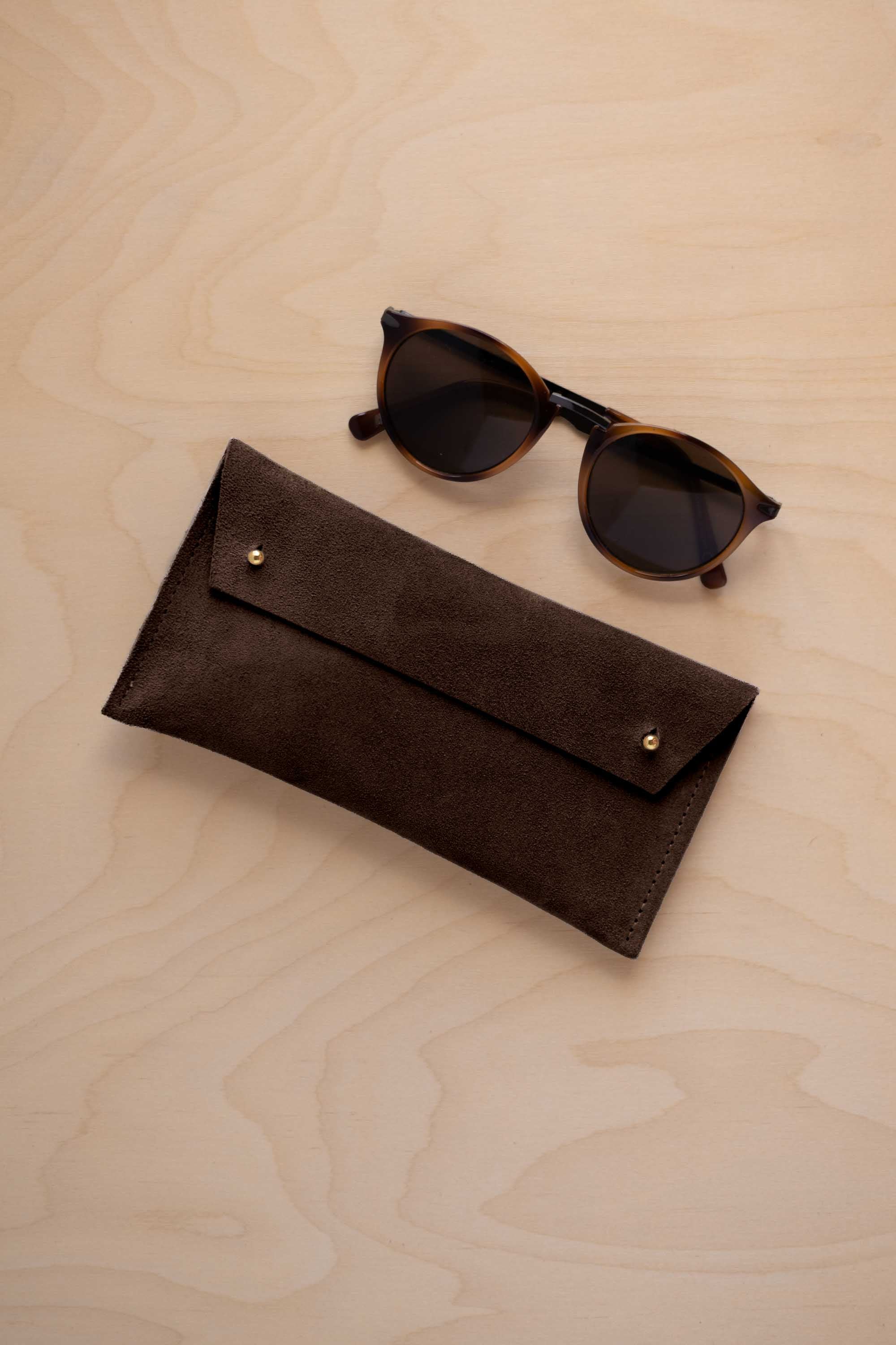 A pair of dark brown tortoiseshell sunglasses with round frames, resting on a light beige wood-grain surface next to an espresso brown coloured suede glasses case with gold coloured buttons.