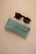 A pair of dark brown tortoiseshell sunglasses with round frames, resting on a light beige wood-grain surface next to a eucalyptus sage green coloured suede glasses case with gold coloured buttons.