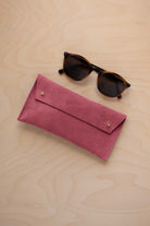 A pair of dark brown tortoiseshell sunglasses with round frames, resting on a light beige wood-grain surface next to a faded rose pink coloured suede glasses case with gold coloured buttons.