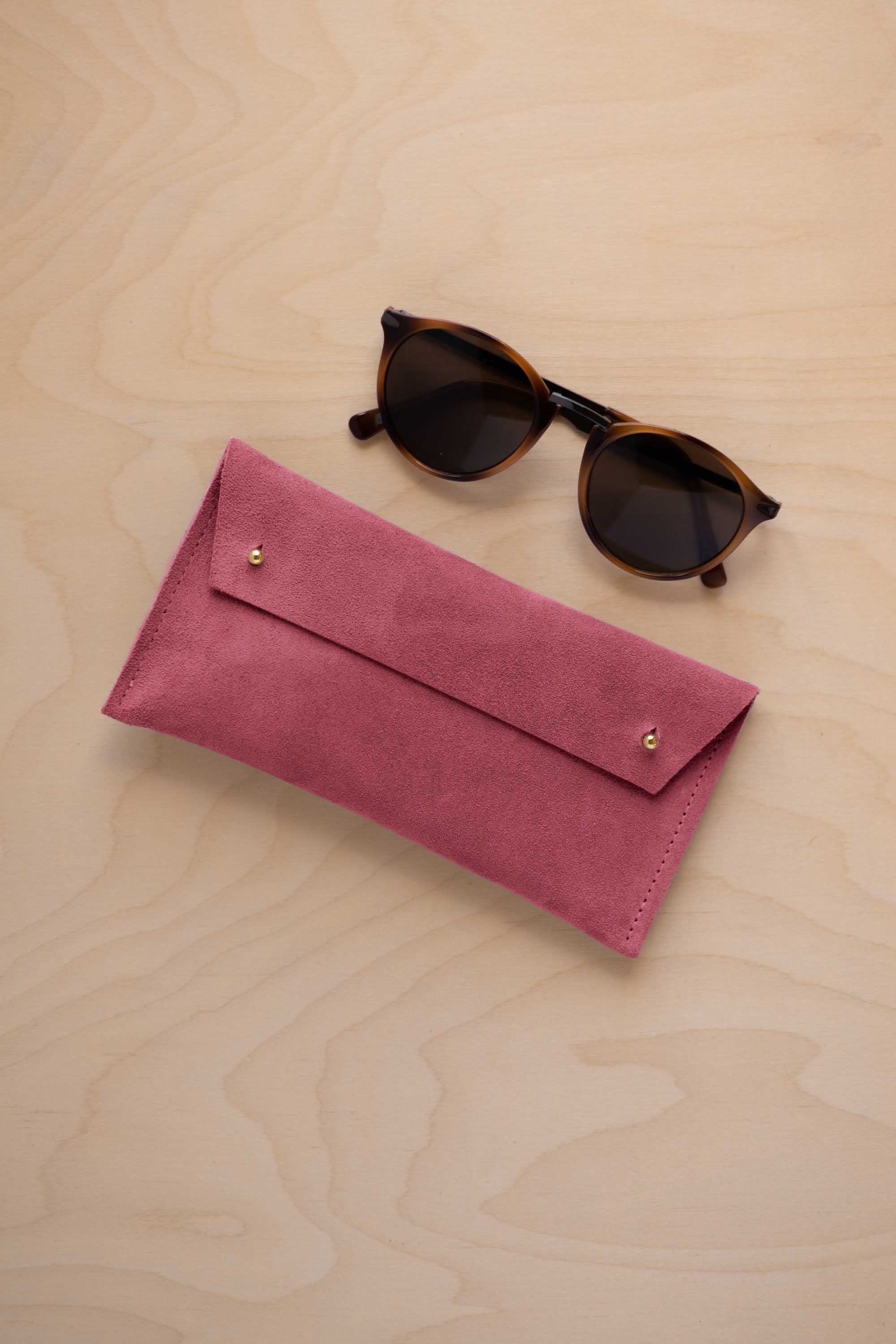 A pair of dark brown tortoiseshell sunglasses with round frames, resting on a light beige wood-grain surface next to a faded rose pink coloured suede glasses case with gold coloured buttons.
