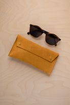 A pair of dark brown tortoiseshell sunglasses with round frames, resting on a light beige wood-grain surface next to a warm beige suede glasses case with gold coloured buttons.