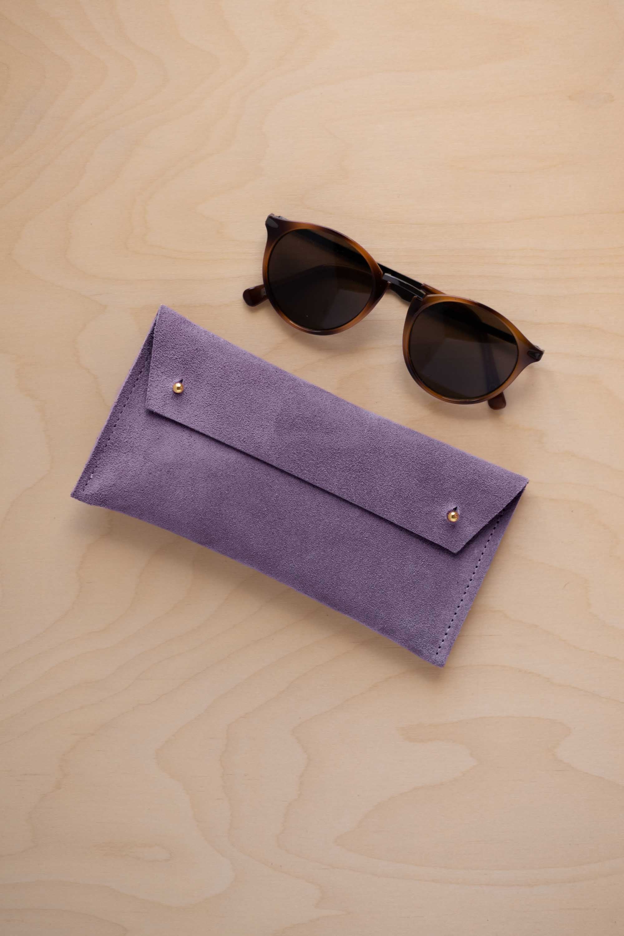 A pair of dark brown tortoiseshell sunglasses with round frames, resting on a light beige wood-grain surface next to a lavender suede glasses case with gold coloured buttons.