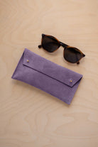 A pair of dark brown tortoiseshell sunglasses with round frames, resting on a light beige wood-grain surface next to a lavender suede glasses case with gold coloured buttons.