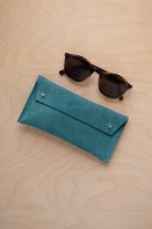A pair of dark brown tortoiseshell sunglasses with round frames, resting on a light beige wood-grain surface next to a marine teal coloured suede glasses case with gold coloured buttons.