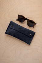 A pair of dark brown tortoiseshell sunglasses with round frames, resting on a light beige wood-grain surface next to a midnight navy blue suede glasses case with gold coloured buttons.