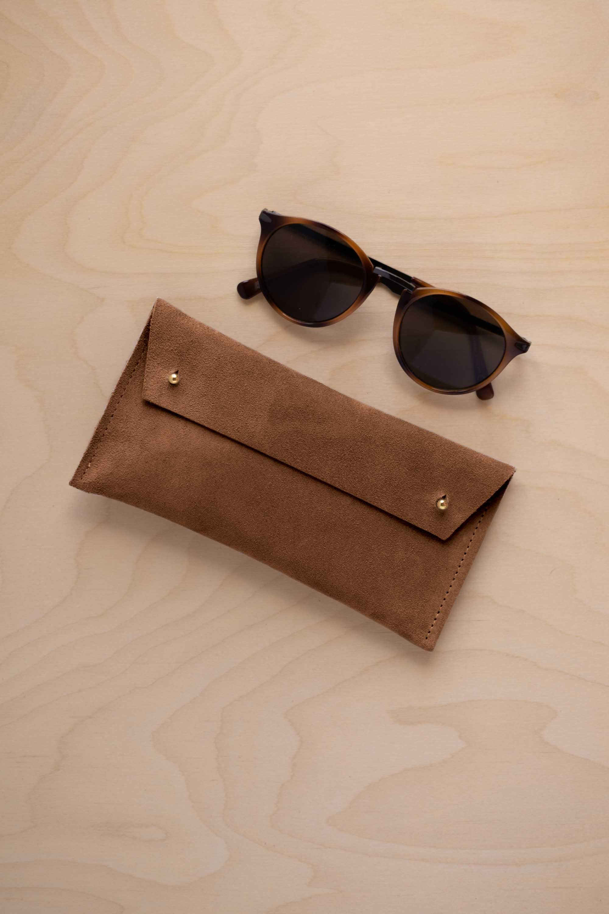 A pair of dark brown tortoiseshell sunglasses with round frames, resting on a light beige wood-grain surface next to a mocha brown suede glasses case with gold coloured buttons.
