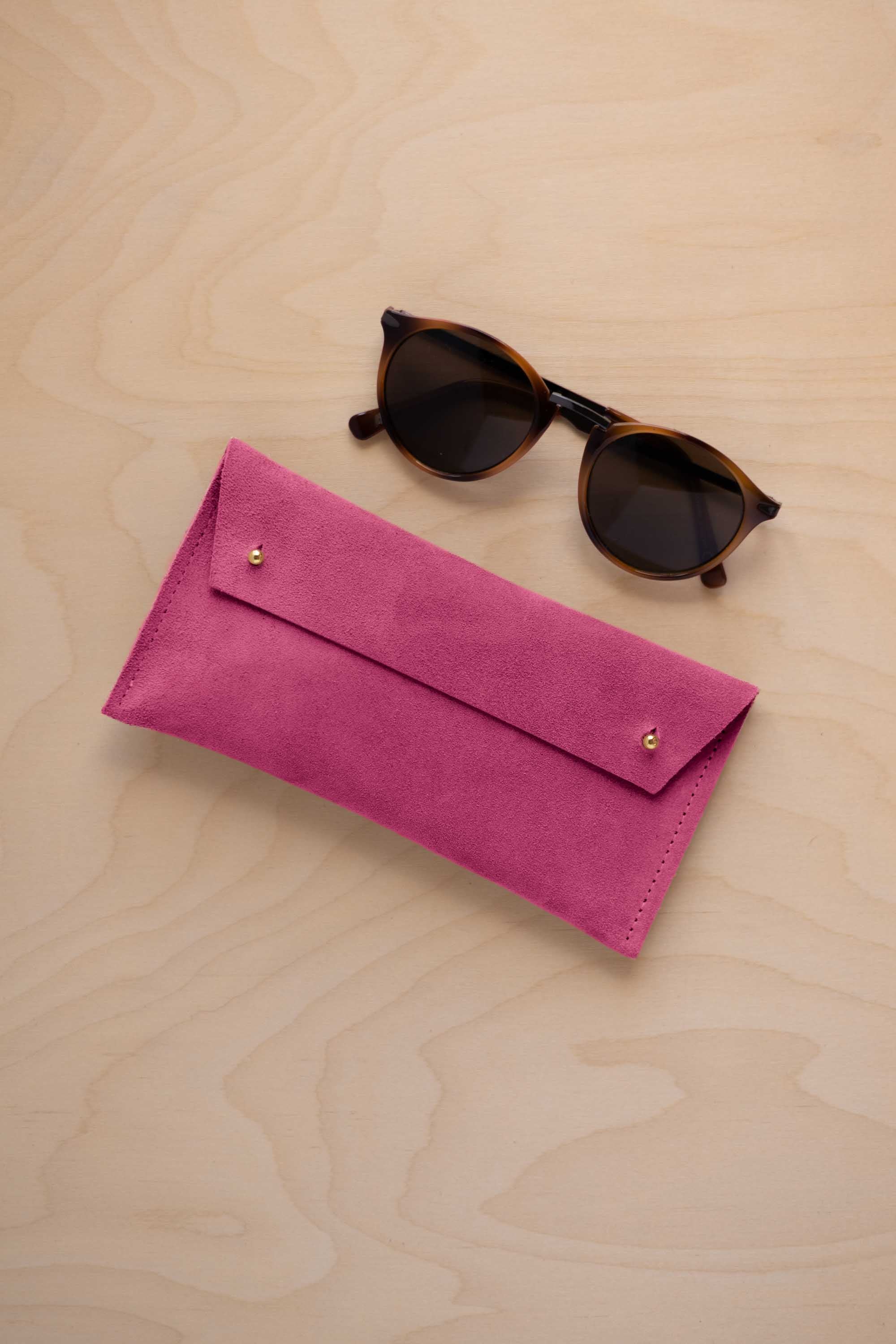 A pair of dark brown tortoiseshell sunglasses with round frames, resting on a light beige wood-grain surface next to a raspberry pink suede glasses case with gold coloured buttons.