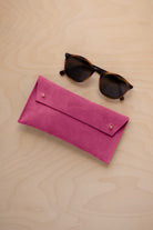 A pair of dark brown tortoiseshell sunglasses with round frames, resting on a light beige wood-grain surface next to a raspberry pink suede glasses case with gold coloured buttons.