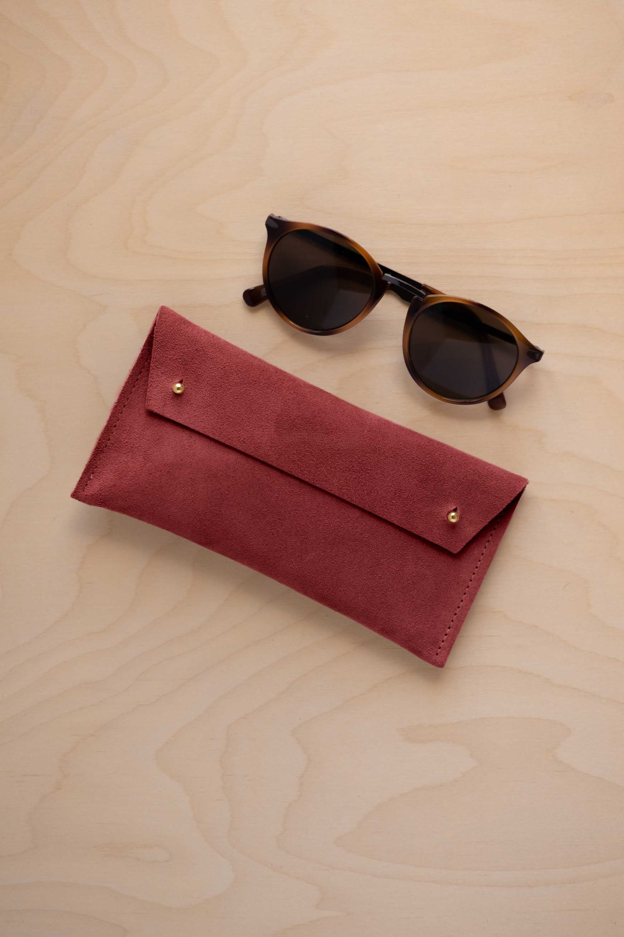 A pair of dark brown tortoiseshell sunglasses with round frames, resting on a light beige wood-grain surface next to a rust red suede glasses case with gold coloured buttons.