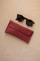 A pair of dark brown tortoiseshell sunglasses with round frames, resting on a light beige wood-grain surface next to a rust red suede glasses case with gold coloured buttons.