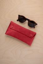 A pair of dark brown tortoiseshell sunglasses with round frames, resting on a light beige wood-grain surface next to a scarlet red suede glasses case with gold coloured buttons.