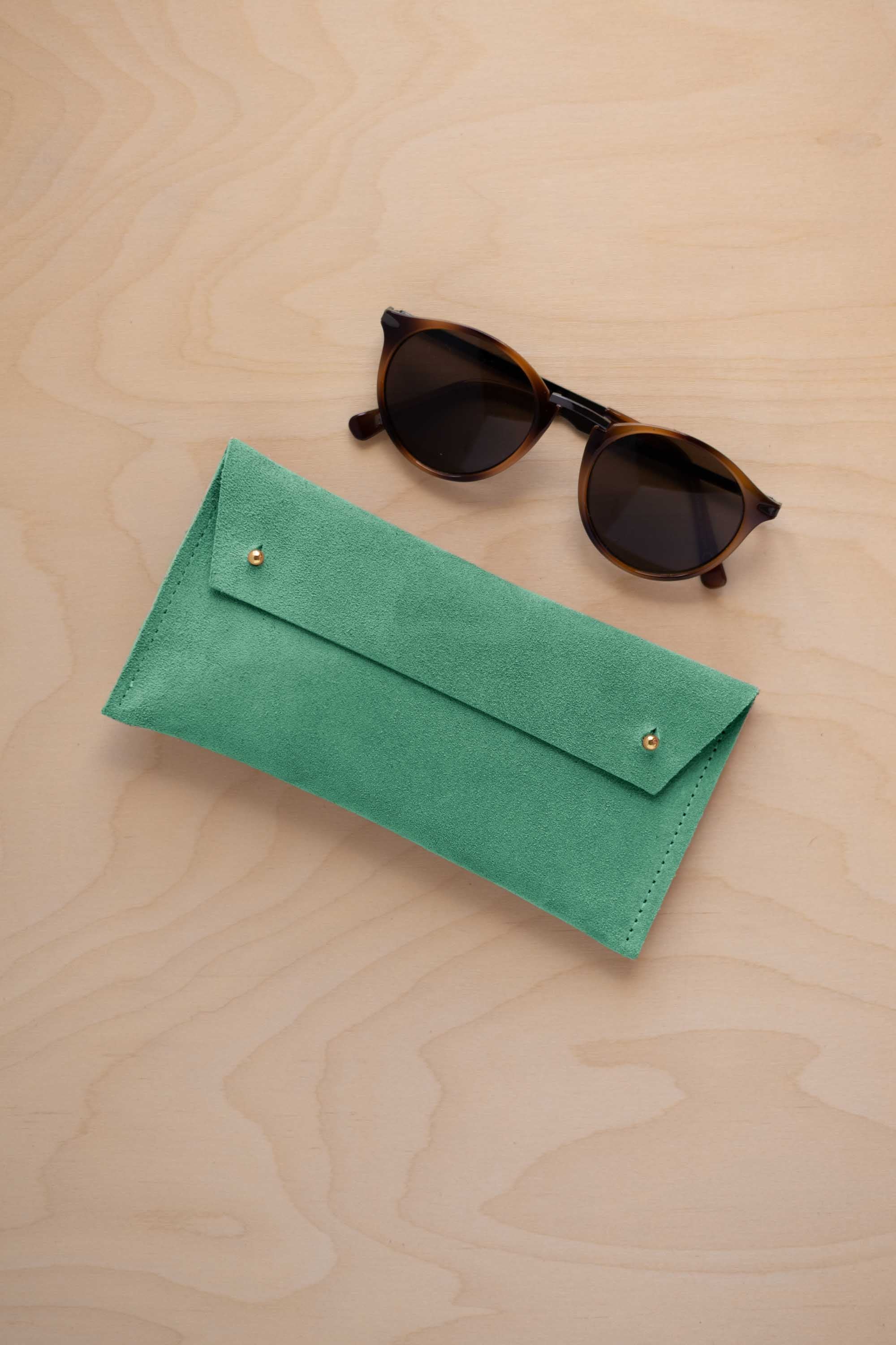 A pair of dark brown tortoiseshell sunglasses with round frames, resting on a light beige wood-grain surface next to a seafoam turquoise green coloured suede glasses case with gold coloured buttons.