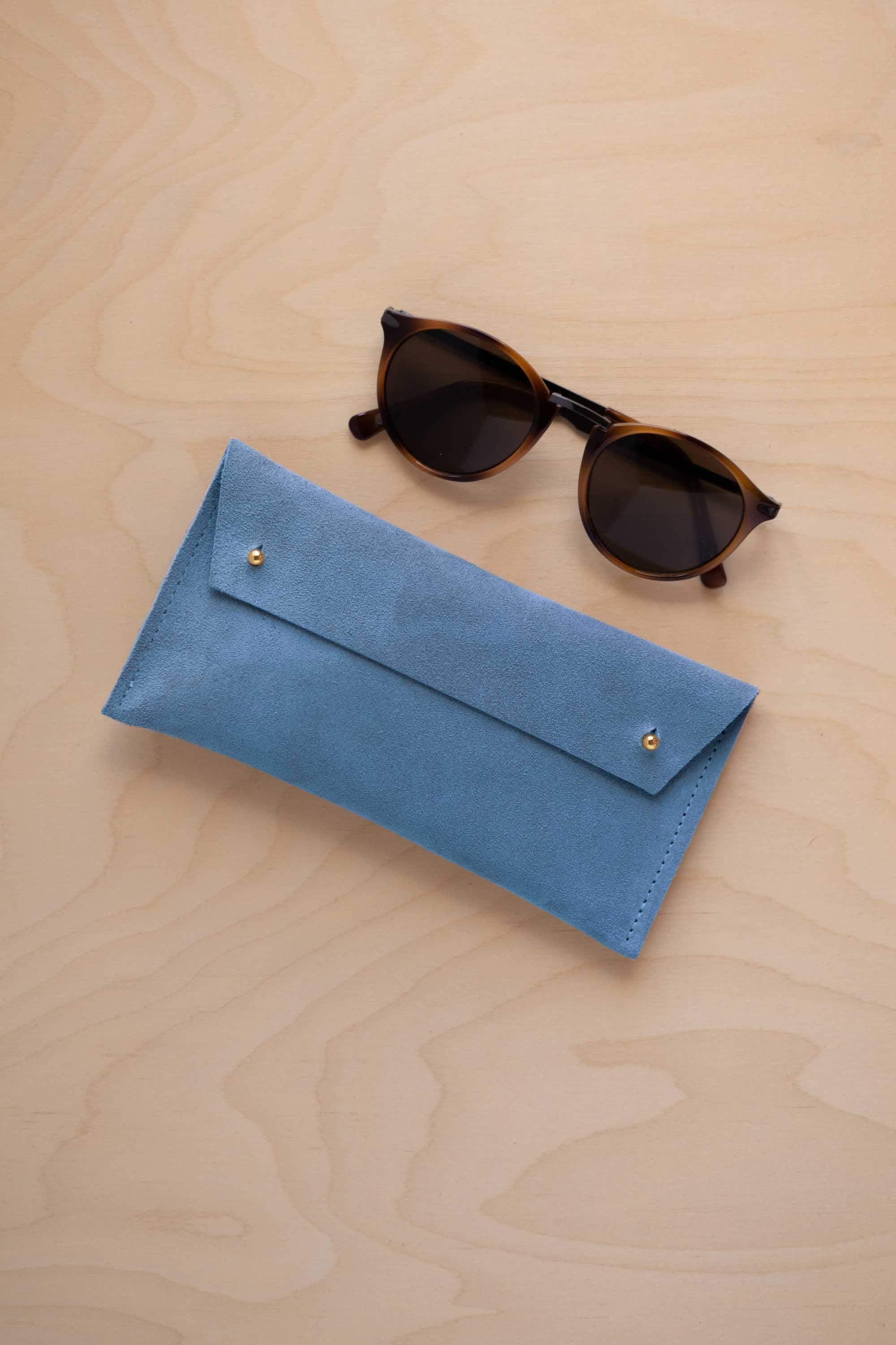 A pair of dark brown tortoiseshell sunglasses with round frames, resting on a light beige wood-grain surface next to a sky blue suede glasses case with gold coloured buttons.