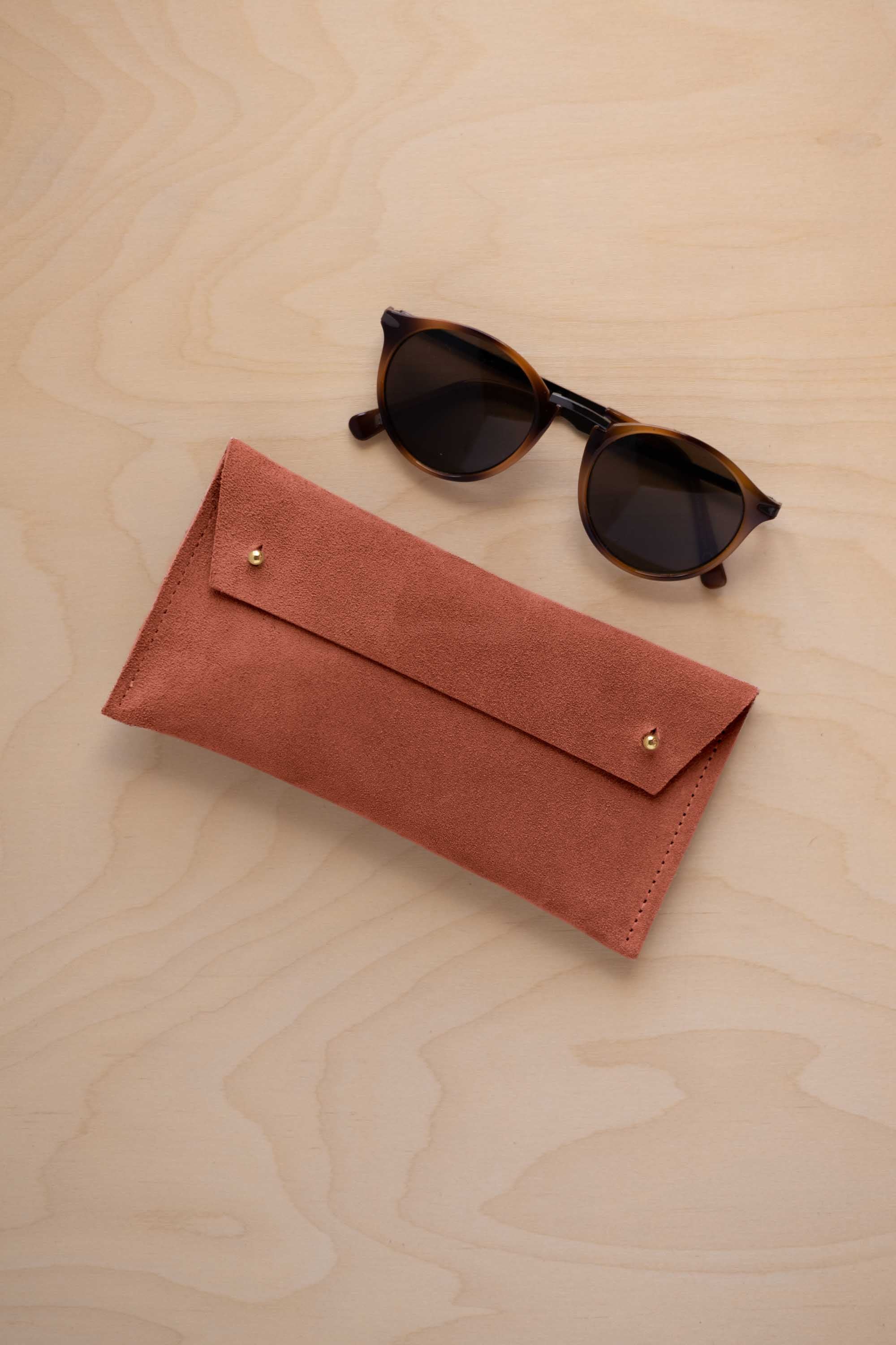 A pair of dark brown tortoiseshell sunglasses with round frames, resting on a light beige wood-grain surface next to a terracotta coloured suede glasses case with gold coloured buttons.