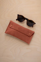 A pair of dark brown tortoiseshell sunglasses with round frames, resting on a light beige wood-grain surface next to a terracotta coloured suede glasses case with gold coloured buttons.