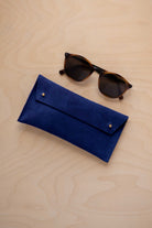 A pair of dark brown tortoiseshell sunglasses with round frames, resting on a light beige wood-grain surface next to an ultramarine blue suede glasses case with gold coloured buttons.