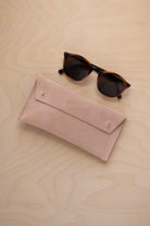 A pair of round sunglasses with dark brown tortoiseshell frames and dark lenses, resting on a light beige wood-grain surface next to a warm beige suede glasses case with gold coloured buttons.