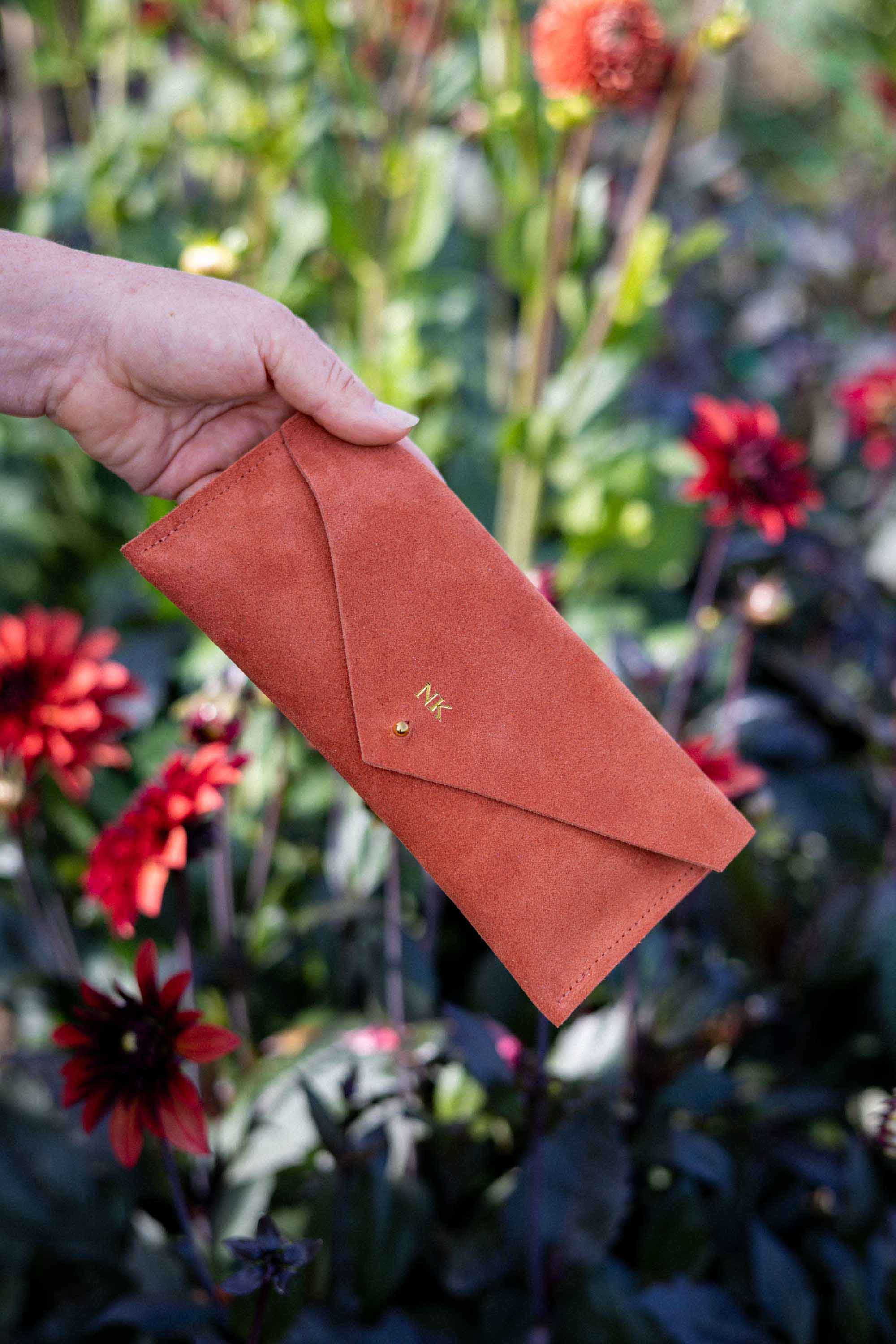 A hand holding a terracotta-colored suede envelope-style wallet or clutch with a small gold monogram, set against a blurred garden background with red dahlia flowers.