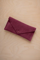 A burgundy suede envelope clutch wallet or bag with a small gold button clasp, resting on a light beige wood grain background.