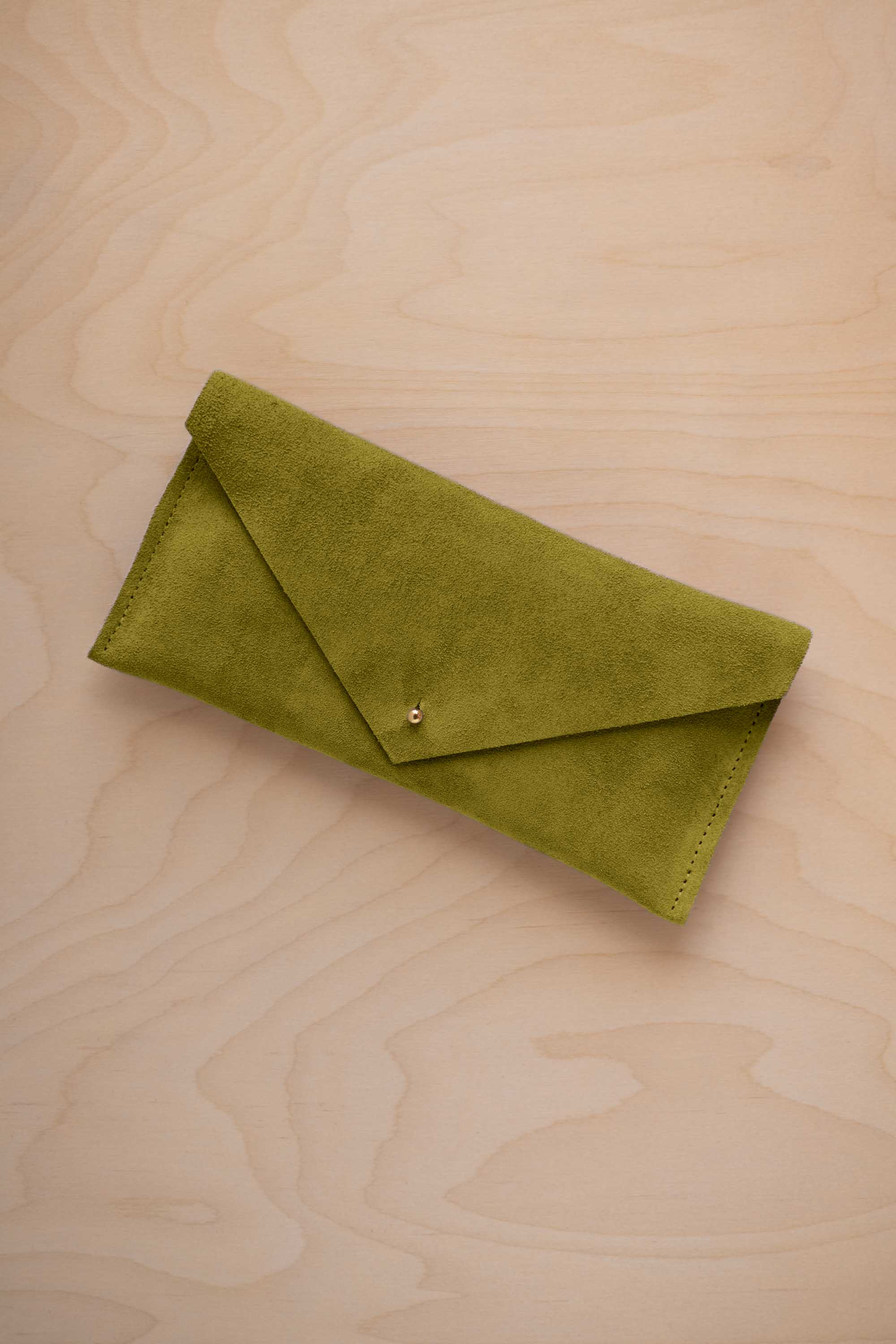 A chartreuse lime green coloured suede envelope clutch wallet or bag with a small gold button clasp, resting on a light beige wood grain background.