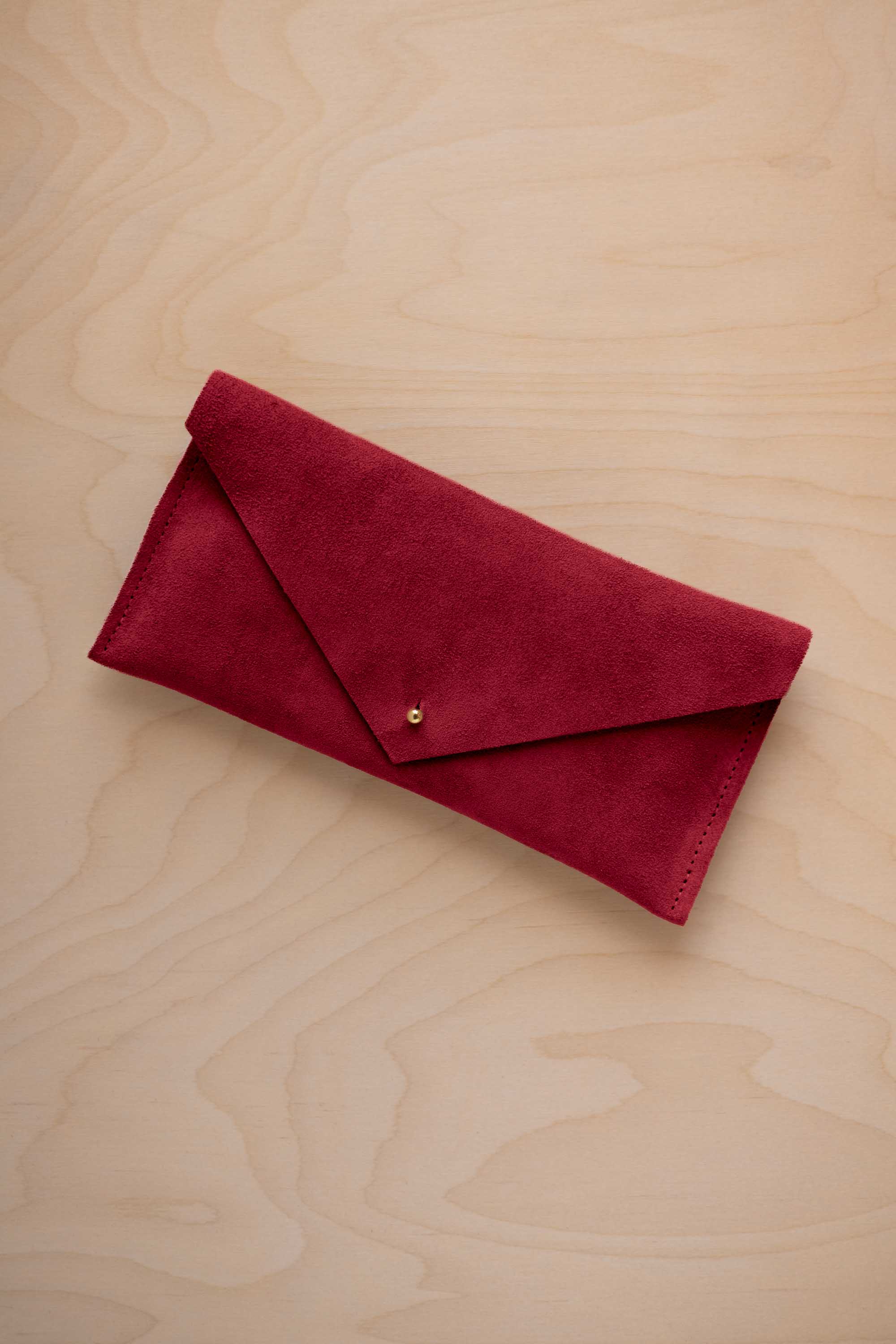 A crimson red suede envelope clutch wallet or bag with a small gold button clasp, resting on a light beige wood grain background.