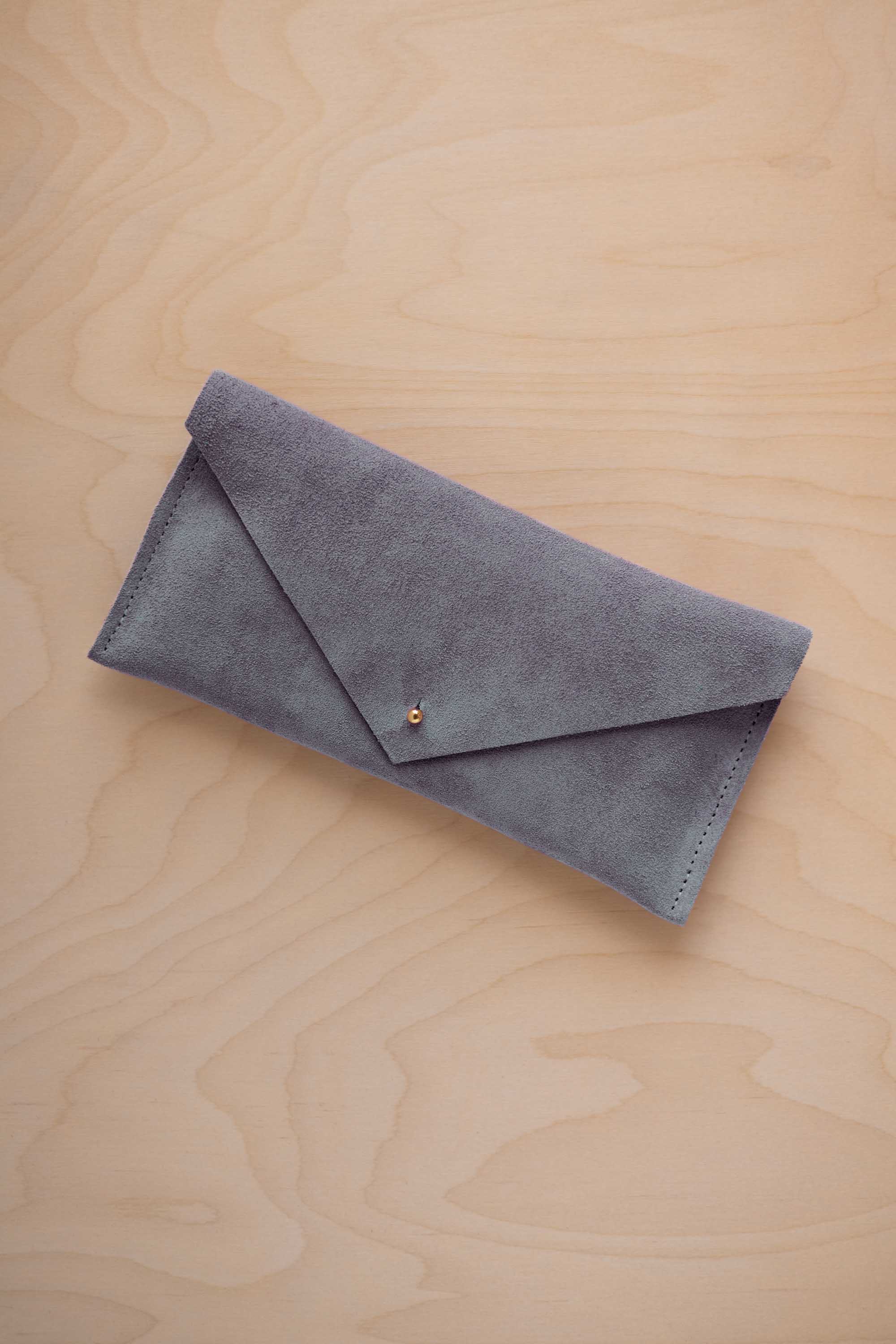 A dusky blue coloured suede envelope clutch wallet or bag with a small gold button clasp, resting on a light beige wood grain background.