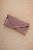 A dusty mauve grey coloured suede envelope clutch wallet or bag with a small gold button clasp, resting on a light beige wood grain background.