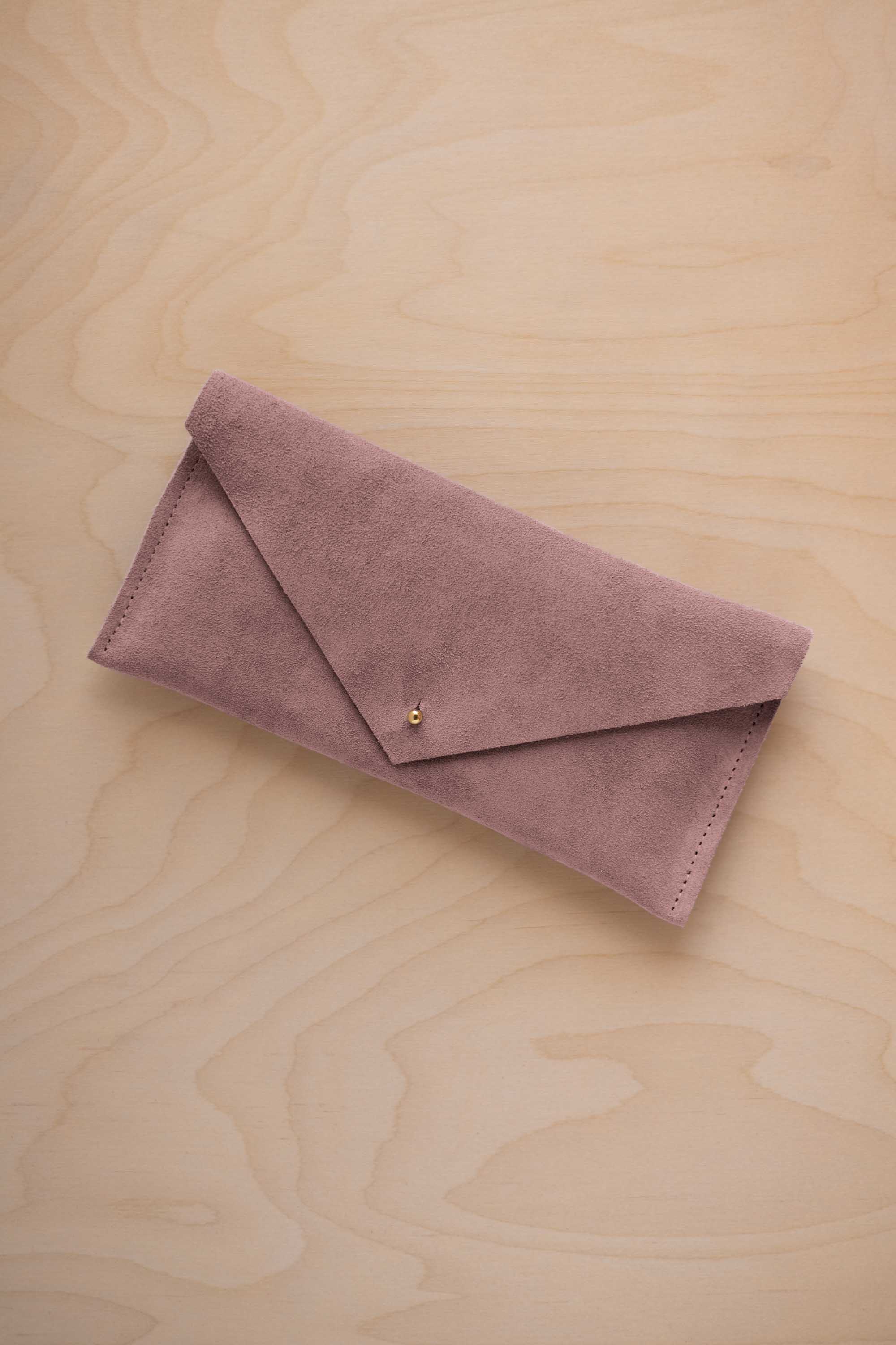A dusty mauve grey coloured suede envelope clutch wallet or bag with a small gold button clasp, resting on a light beige wood grain background.