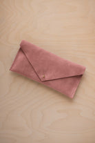 A dusty rose pink suede envelope clutch wallet or bag with a small gold button clasp, resting on a light beige wood grain background.