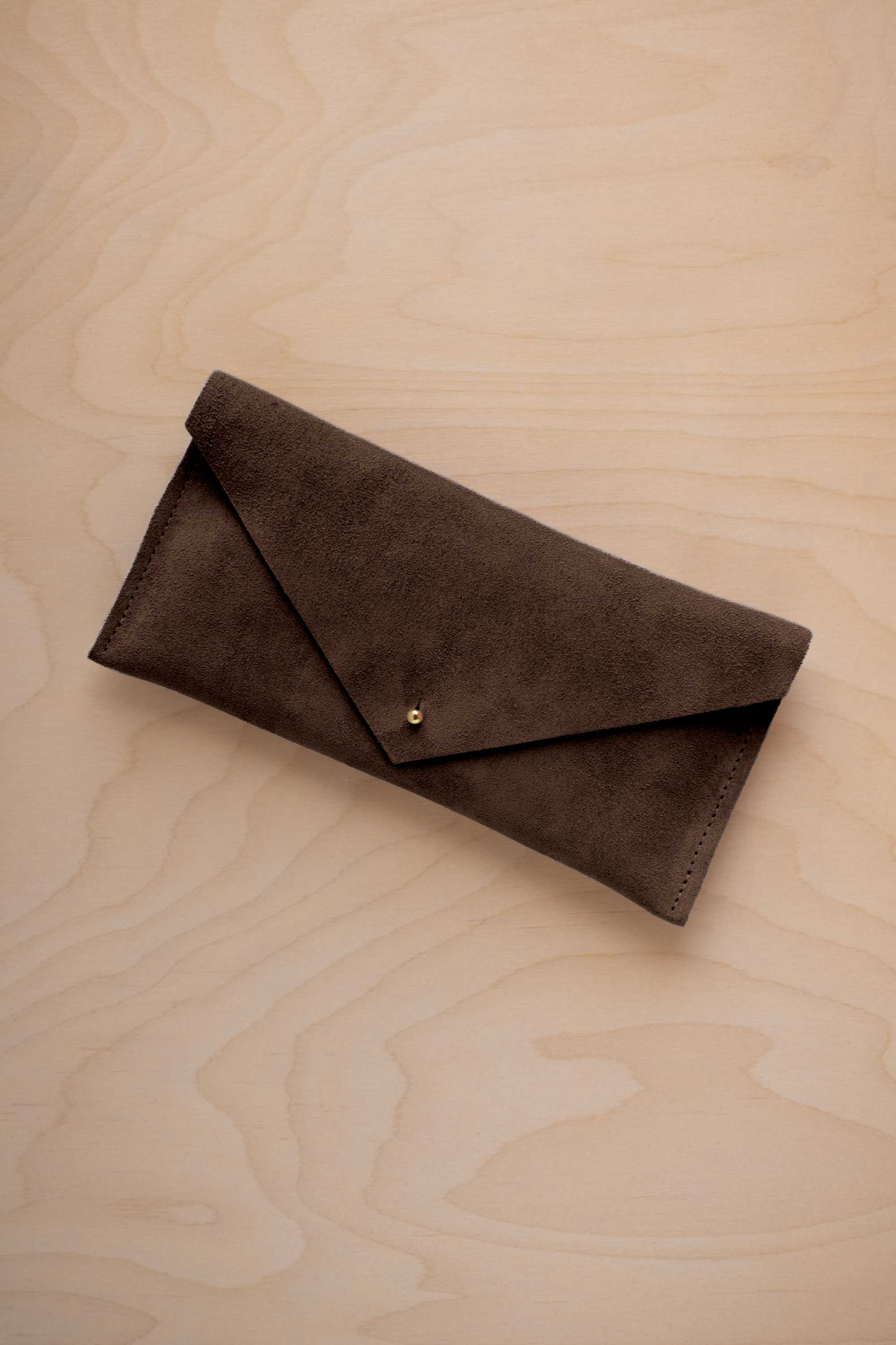 An espresso brown suede envelope clutch wallet or bag with a small gold button clasp, resting on a light beige wood grain background.