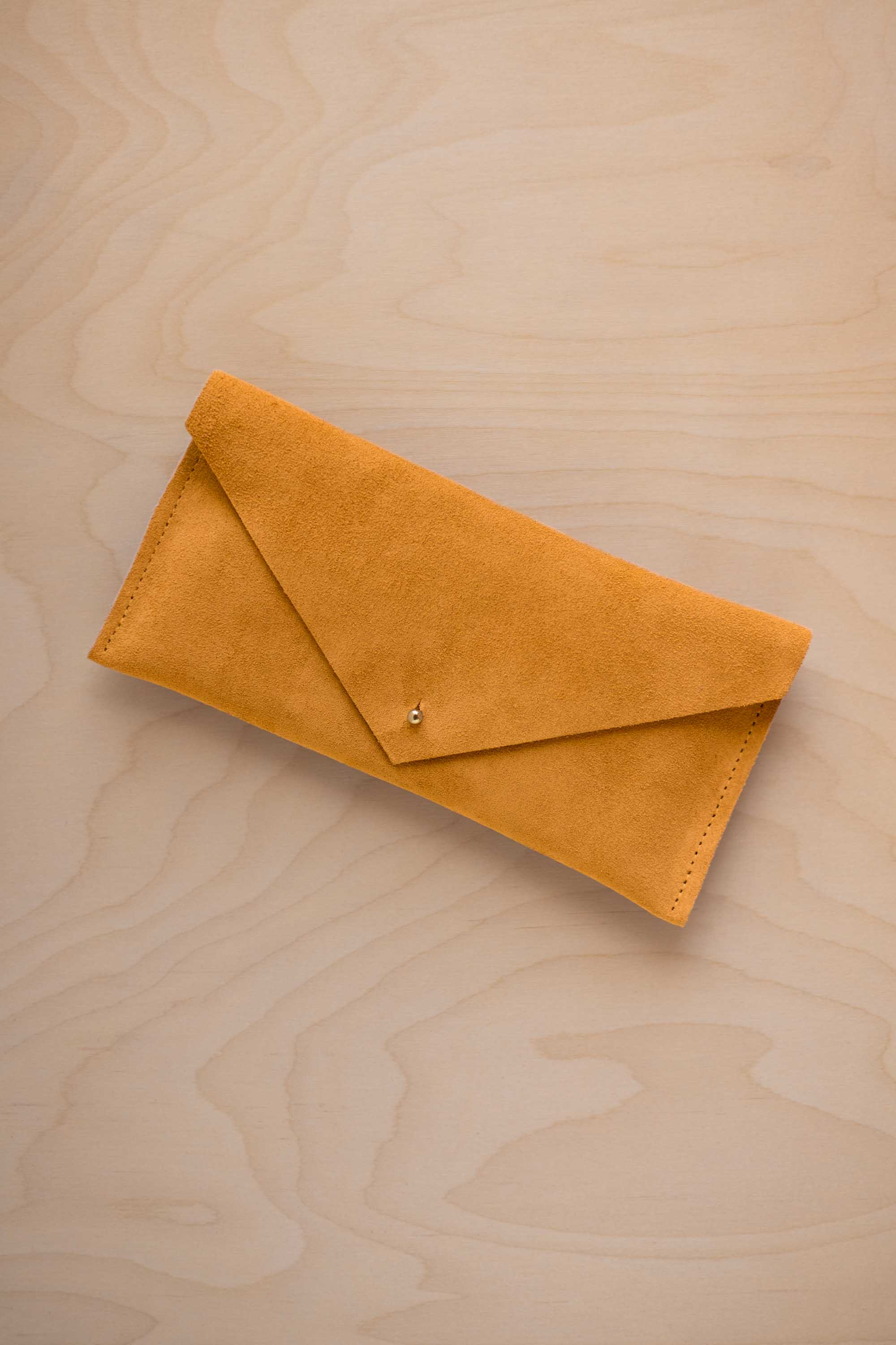 A golden ochre mustard yellow coloured envelope clutch wallet or bag with a small gold button clasp, resting on a light beige wood grain background.