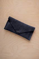 A midnight navy blue coloured suede envelope clutch wallet or bag with a small gold button clasp, resting on a light beige wood grain background.