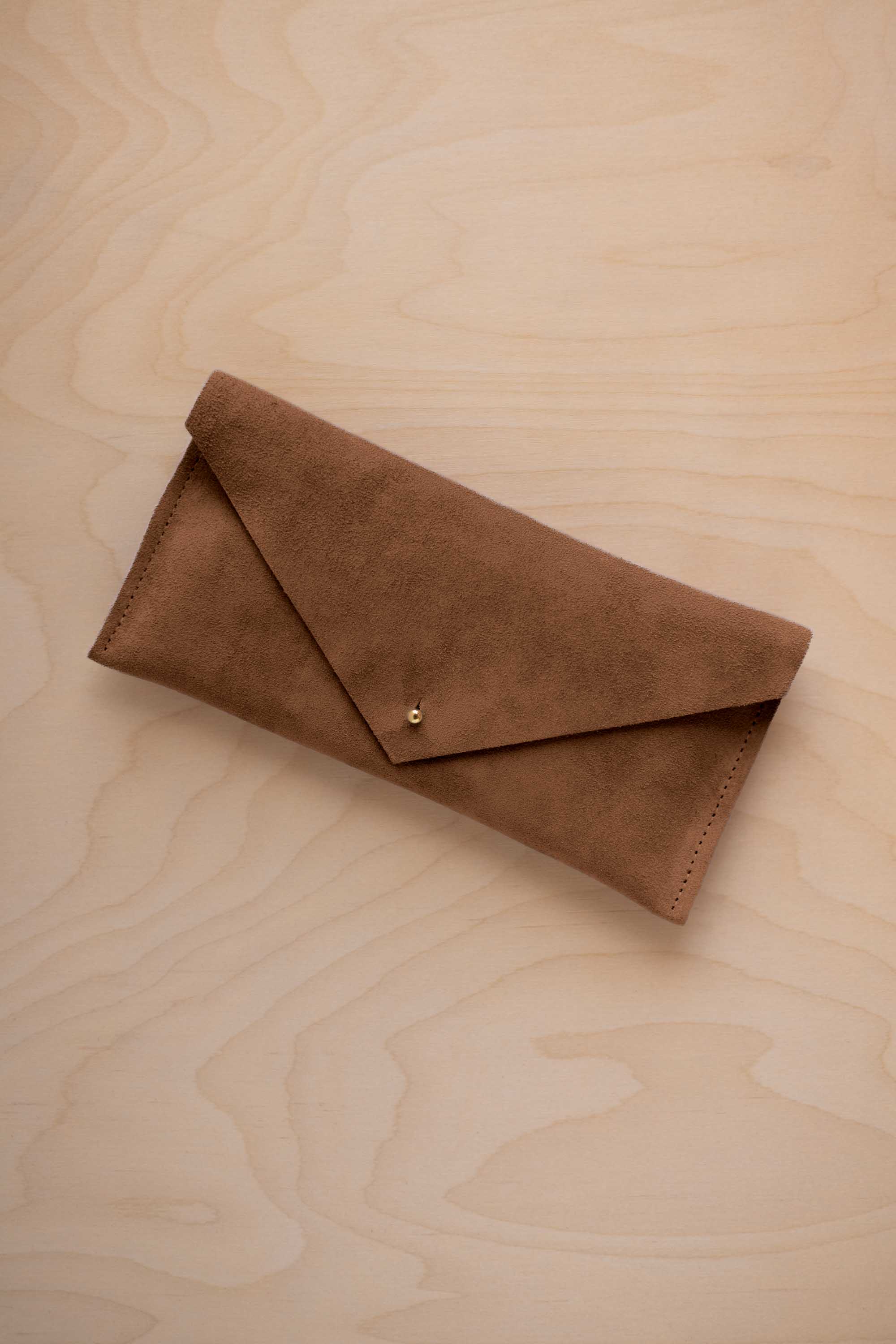 A mocha brown envelope clutch wallet or bag with a small gold button clasp, resting on a light beige wood grain background.