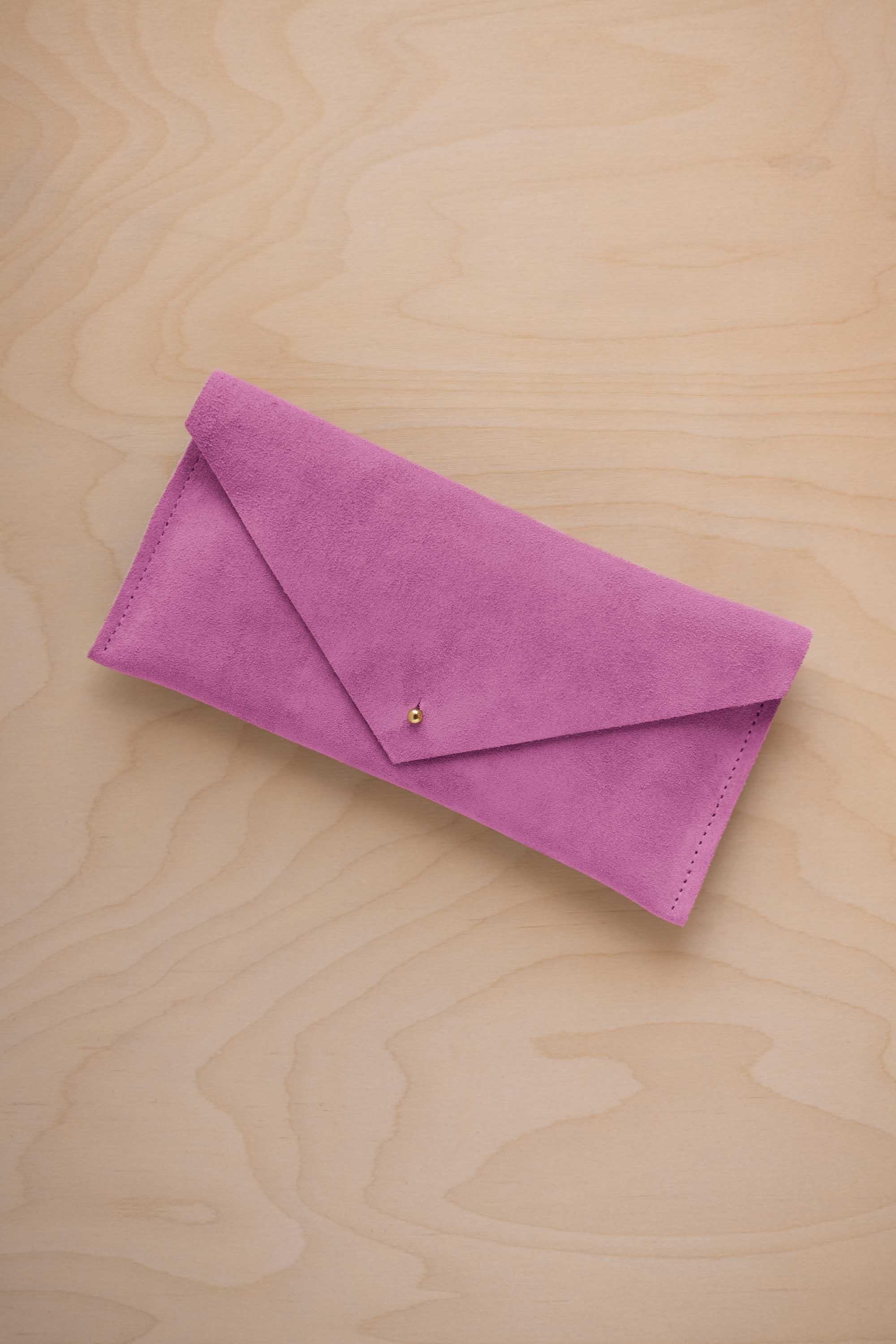 A peony pink coloured suede envelope clutch wallet or bag with a small gold button clasp, resting on a light beige wood grain background.