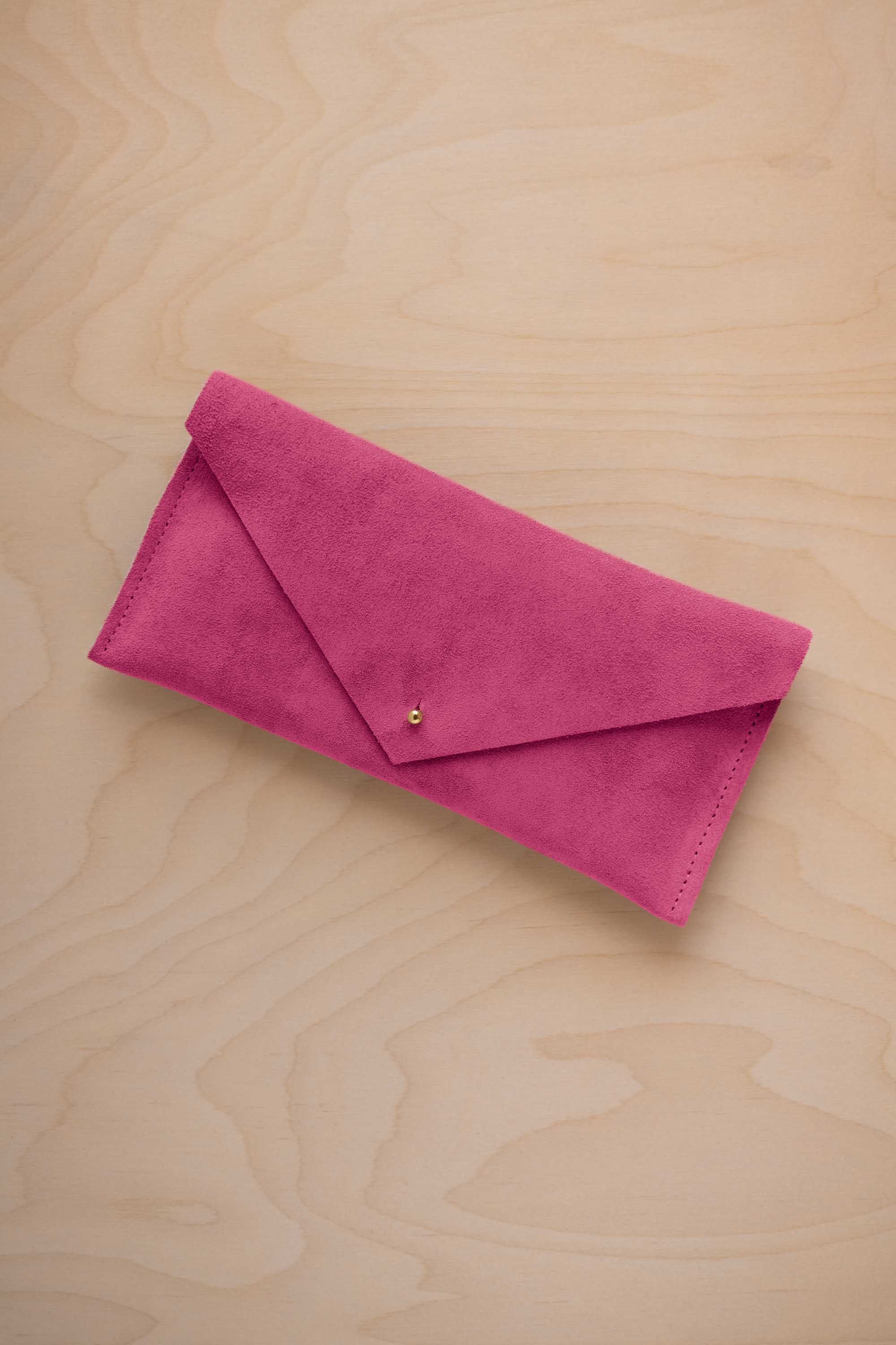 A raspberry pink coloured suede envelope clutch wallet or bag with a small gold button clasp, resting on a light beige wood grain background.