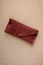 A rust red coloured suede envelope clutch wallet or bag with a small gold button clasp, resting on a light beige wood grain background.